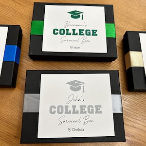 May include: Black gift boxes with white labels and ribbon accents. The labels read "College Survival Box" with a graduation cap graphic and a name. One box has green ribbon, another has silver, and a third has blue.