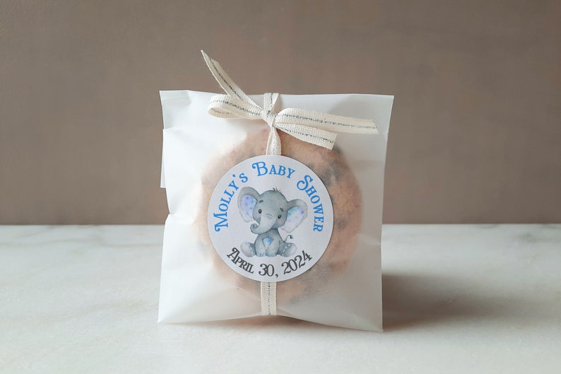 Pu&ograve; includere: Un sacchetto regalo bianco con un adesivo rotondo blu e bianco che dice "Baby Shower di Molly" con un elefante dei cartoni animati e la data "30 aprile 2024".