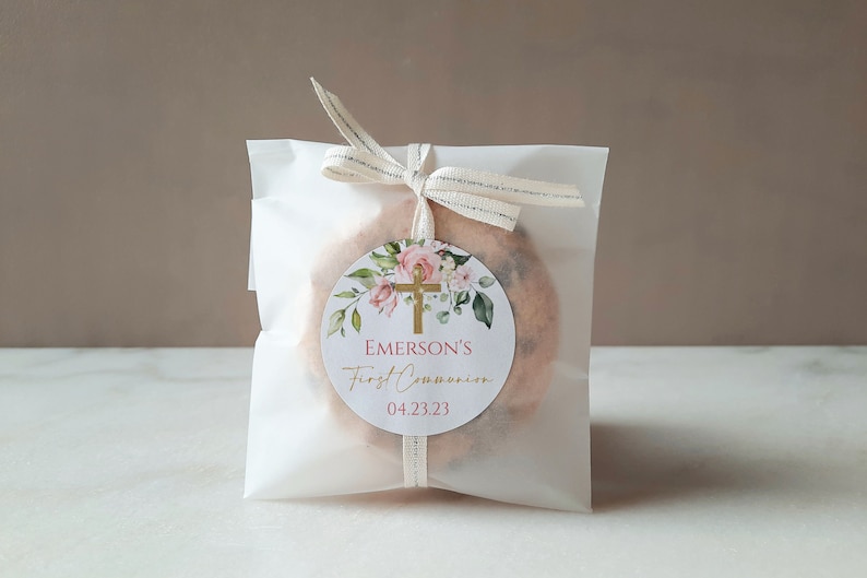 May include: A white favor bag with a gold cross and pink floral design. The bag is tied with a white ribbon and has a personalized label that reads "Emerson's First Communion 04.23.23".