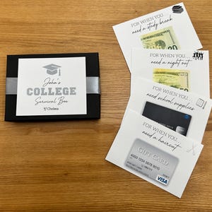 May include: A black box with a white label that reads "John's COLLEGE Survival Box" with a graduation cap graphic. Several white cards with text and money or gift cards are next to the box. The cards say "For when you need a study break", "For when you need a night out", "For when you need school supplies", and "For when you need a haircut".