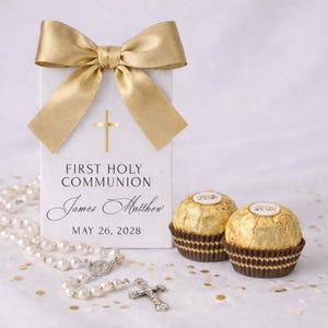 May include: A white gift box with a gold bow, cross, and the words "First Holy Communion", a name and date. A rosary and two gold-wrapped chocolates are also present. The theme is religious and celebratory.