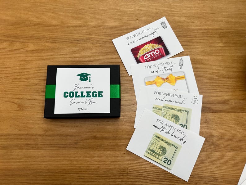 May include: A black box with a green ribbon, labeled "Breanna's College Survival Box." Includes gift cards for a movie night and a treat, cash, and a gift card for laundry. The box is on a wooden surface.