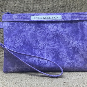 May include: Purple zippered pouch with a white strap and a white label that reads "EVEN KEEL & CO". The pouch is decorated with a white dragonfly pattern.