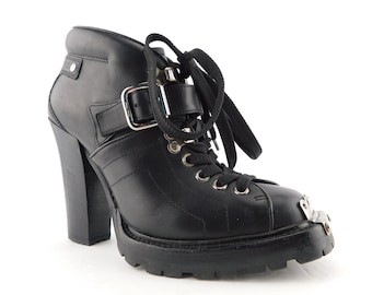 May include: A black leather ankle boot with a high block heel, a thick lug sole, and black laces. The boot features a silver-toned metal toe cap, a side strap with a silver buckle, and silver eyelets for the laces, creating a structured look.