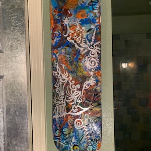Può includere: Un mazzo di skateboard dipinto con arte astratto in tonalità di blu, arancione e bianco. L'opera d'arte presenta motivi a spirale e un viso stilizzato.