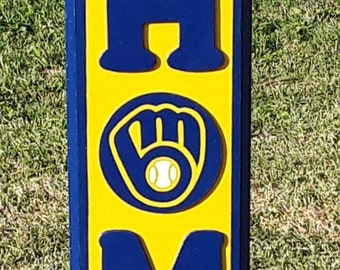 Könnte beinhalten: Ein gelbes und blaues Holzschild mit dem Wort "HOME" in großen blauen Buchstaben. Das "M" wird durch das Logo der Milwaukee Brewers ersetzt, ein blauer Handschuh mit einem weißen Baseball. Das Schild trägt den Text "MILWAUKEE BREWERS "HOME" SIGN" und "LET EVERYONE KNOW YOUR HOME IS A "BREW CREW" HOME WITH THIS AWESOME SIGN!"