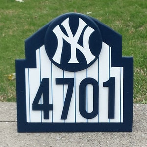 May include: A blue and white wooden sign with a New York Yankees logo and the number 4701. The sign is shaped like a baseball stadium.