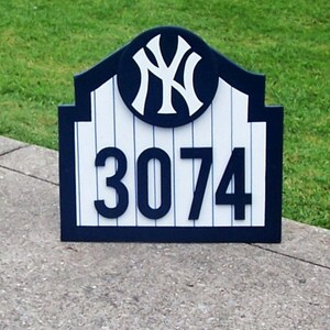 May include: A blue and white wooden sign with a New York Yankees logo and the number 3074. The sign is shaped like a baseball stadium arch.