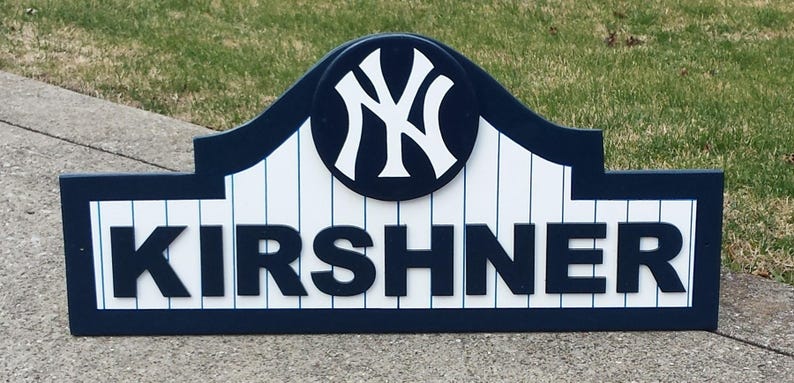 Pu&ograve; includere: Un cartello di legno con sfondo a strisce blu e bianche. Il cartello presenta un logo bianco dei New York Yankees in un cerchio blu e il nome "KIRSHNER" in lettere blu.