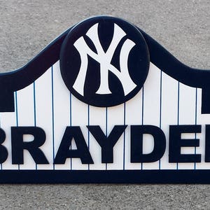 Pu&ograve; includere: Un cartello in legno blu scuro e bianco con un design a tema baseball. Il cartello presenta un logo bianco dei New York Yankees in un cerchio e il nome "BRAYDEN" in grandi lettere nere.