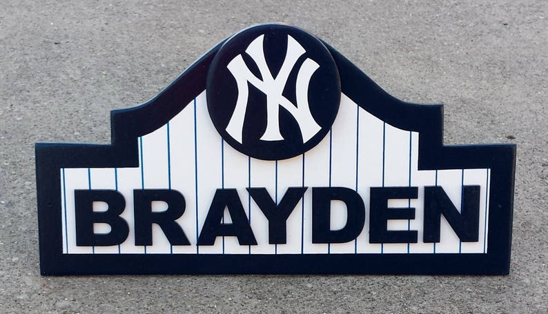 Pu&ograve; includere: Un cartello in legno blu scuro e bianco con un design a tema baseball. Il cartello presenta un logo bianco dei New York Yankees in un cerchio e il nome "BRAYDEN" in grandi lettere nere.