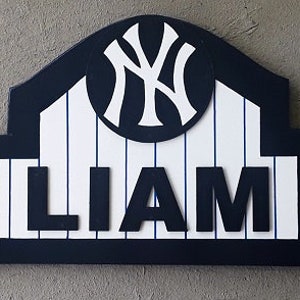 Pu&ograve; includere: Un cartello in legno con un tema da baseball blu e bianco. Il cartello presenta il logo dei New York Yankees e il nome "LIAM" in lettere bianche.