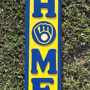 Op de afbeelding: Een geel en blauw houten bord met het woord "HOME" in blauwe letters. De "O" is vervangen door een blauwe honkbalhandschoen met een witte honkbal.