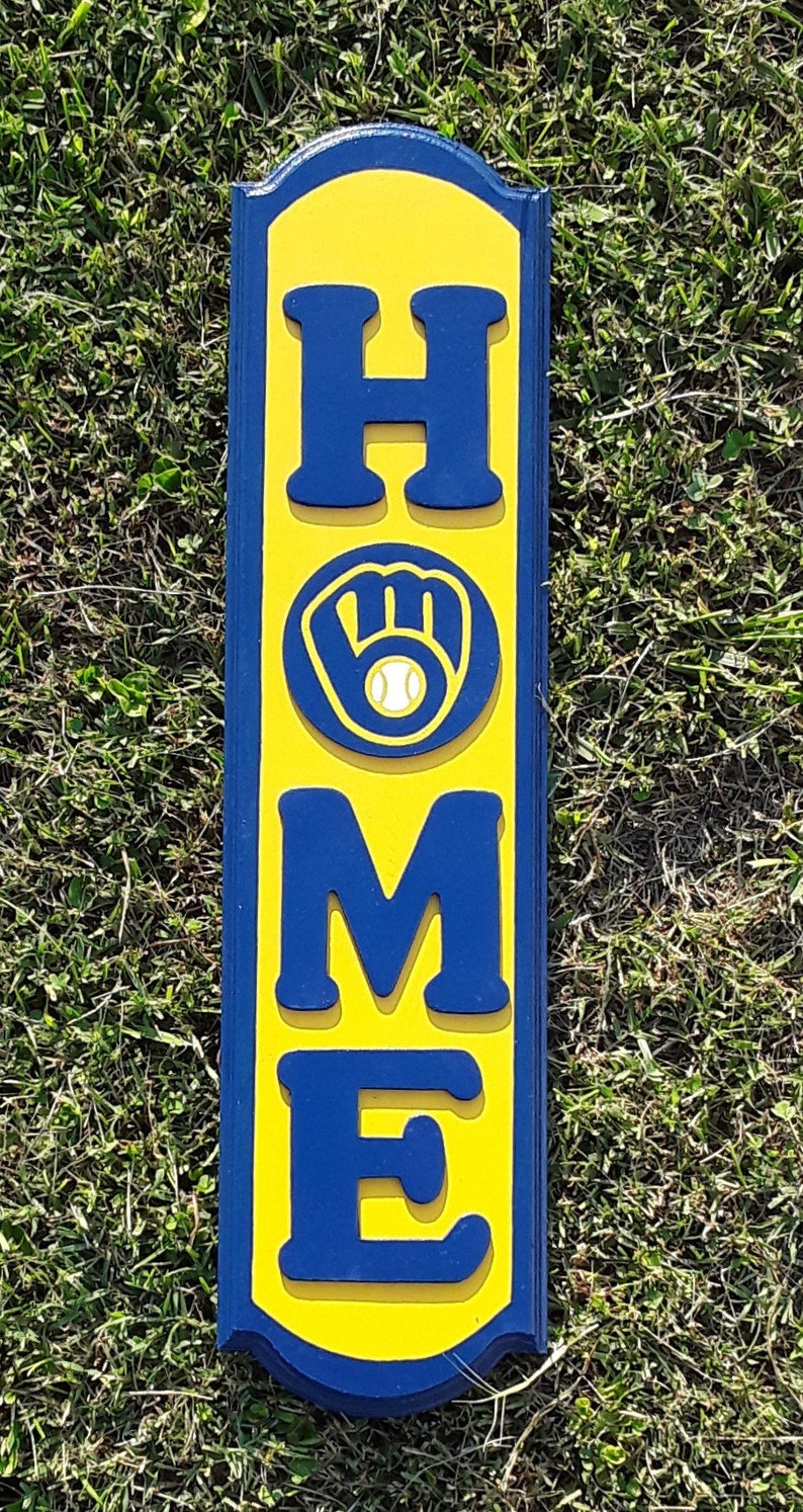 Op de afbeelding: Een geel en blauw houten bord met het woord "HOME" in blauwe letters. De "O" is vervangen door een blauwe honkbalhandschoen met een witte honkbal.