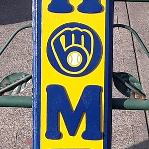 Op de afbeelding: Een houten bord met het woord "HOME" in blauwe letters op een gele achtergrond. De letter "O" is vervangen door een blauwe honkbalhandschoen met een witte honkbal erin.
