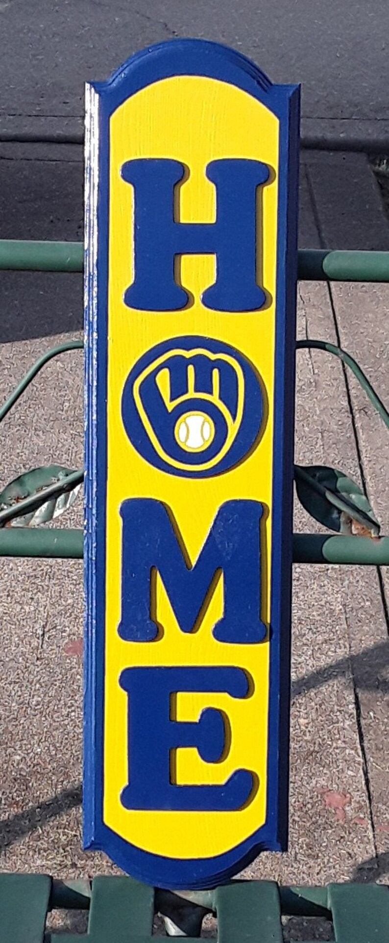 Op de afbeelding: Een houten bord met het woord "HOME" in blauwe letters op een gele achtergrond. De letter "O" is vervangen door een blauwe honkbalhandschoen met een witte honkbal erin.