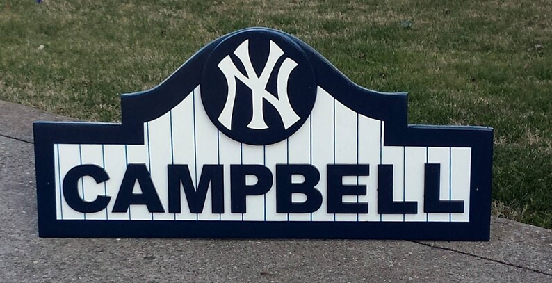 Pu&ograve; includere: Un cartello in legno con un design a tema baseball blu e bianco. Il cartello presenta un logo bianco dei New York Yankees con un contorno blu e il nome "CAMPBELL" in lettere blu.