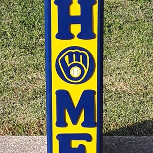 Op de afbeelding: Een houten bord met het woord "HOME" gespeld in blauwe letters op een gele achtergrond. Een blauwe honkbalhandschoen met een witte honkbal bevindt zich in het midden van het bord.