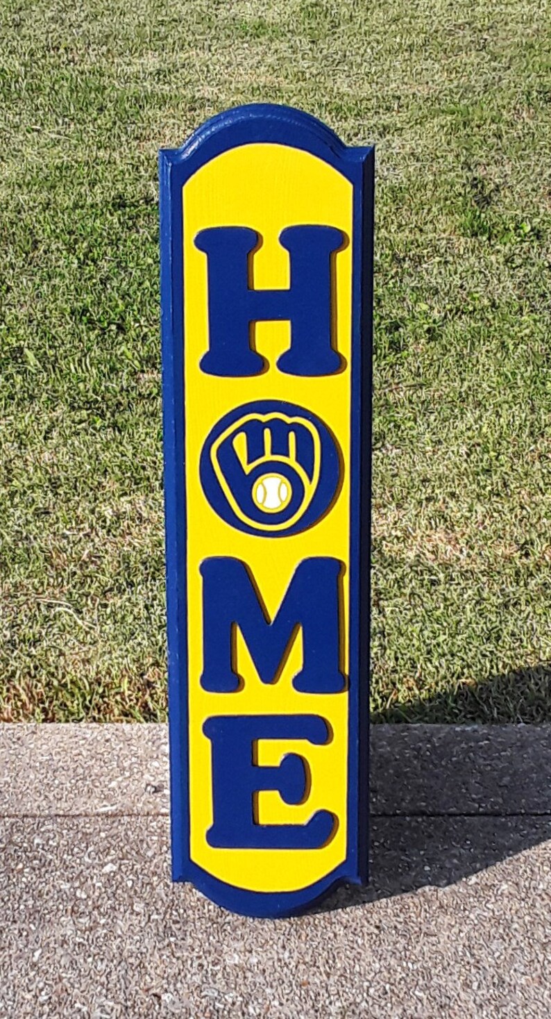 Op de afbeelding: Een houten bord met het woord "HOME" gespeld in blauwe letters op een gele achtergrond. Een blauwe honkbalhandschoen met een witte honkbal bevindt zich in het midden van het bord.