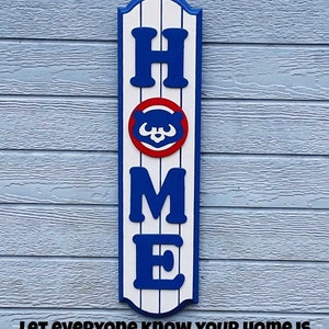 May include: A wooden sign with the word "HOME" in blue letters. The "O" is replaced with the Chicago Cubs logo. The sign has a white and blue striped background. The text "Let everyone know your home is a "CUBS HOME" is below the sign.