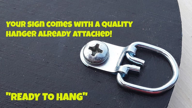 Op de afbeelding: Een close-up van een zwart houten bord met een zilveren metalen hanger eraan bevestigd. Het bord heeft de tekst "YOUR SIGN COMES WITH A QUALITY HANGER ALREADY ATTACHED!" en "READY TO HANG" in gele letters.