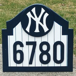 May include: A blue and white wooden sign with a New York Yankees logo and the house number 6780. The sign is shaped like a baseball stadium.