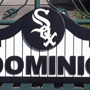 Op de afbeelding: Een zwart-wit bord met de naam "DOMINIC". Het bord toont het logo van de Chicago White Sox in een cirkelvormig embleem. Het bord is gemonteerd op een groene metalen structuur.