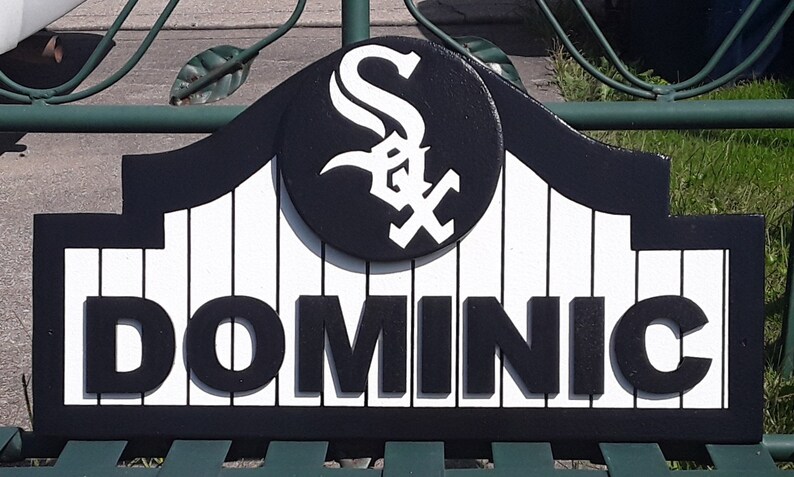 Op de afbeelding: Een zwart-wit bord met de naam "DOMINIC". Het bord toont het logo van de Chicago White Sox in een cirkelvormig embleem. Het bord is gemonteerd op een groene metalen structuur.