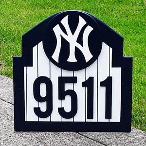 May include: A black and white wooden sign with a New York Yankees logo and the number 9511. The sign is shaped like a baseball stadium and has a white and blue striped background.