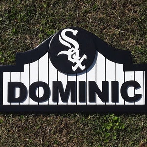 Op de afbeelding: Een gepersonaliseerd houten bord met de naam "DOMINIC" in vette, zwarte letters. Het bord heeft een zwart-wit honkbalteamlogo en een zwarte rand. De achtergrond is wit met verticale zwarte lijnen.