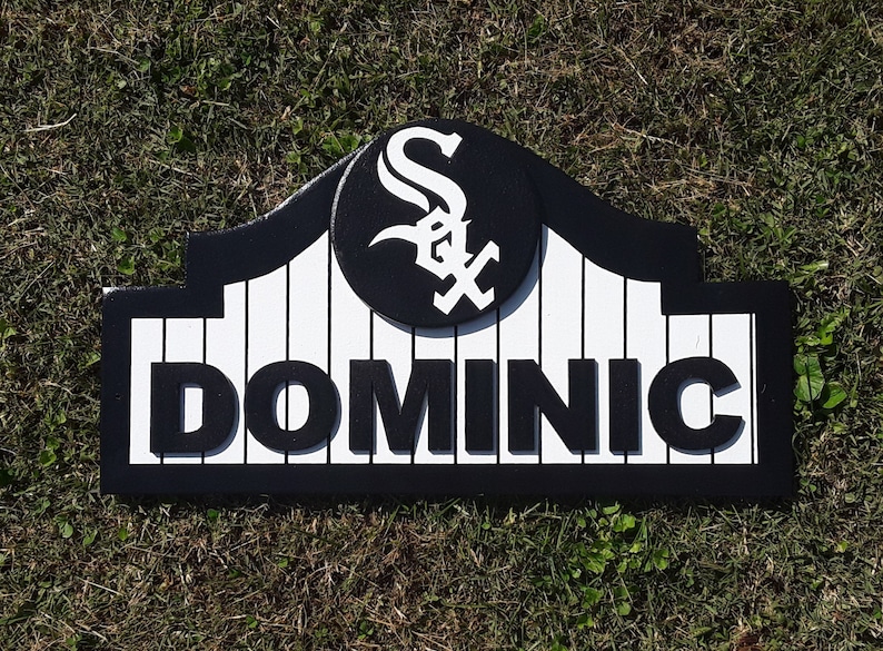 Op de afbeelding: Een gepersonaliseerd houten bord met de naam "DOMINIC" in vette, zwarte letters. Het bord heeft een zwart-wit honkbalteamlogo en een zwarte rand. De achtergrond is wit met verticale zwarte lijnen.