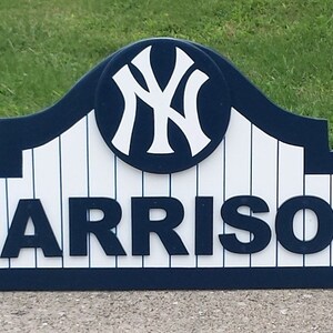 Pu&ograve; includere: Un cartello in legno blu e bianco con il logo dei New York Yankees e il nome "HARRISON" in lettere bianche.