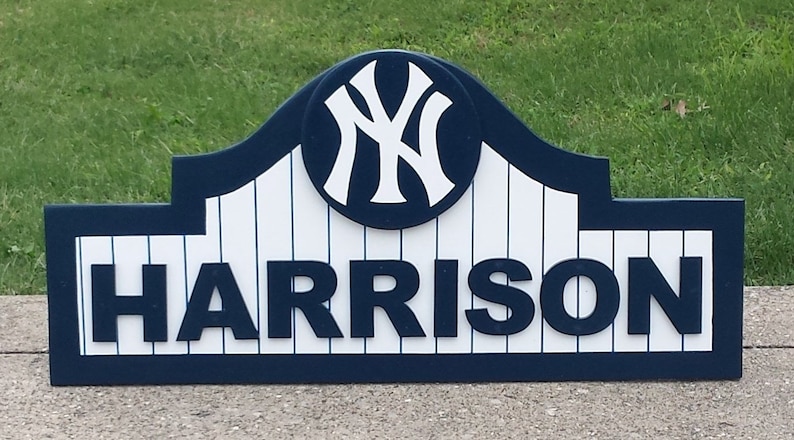 Pu&ograve; includere: Un cartello in legno blu e bianco con il logo dei New York Yankees e il nome "HARRISON" in lettere bianche.
