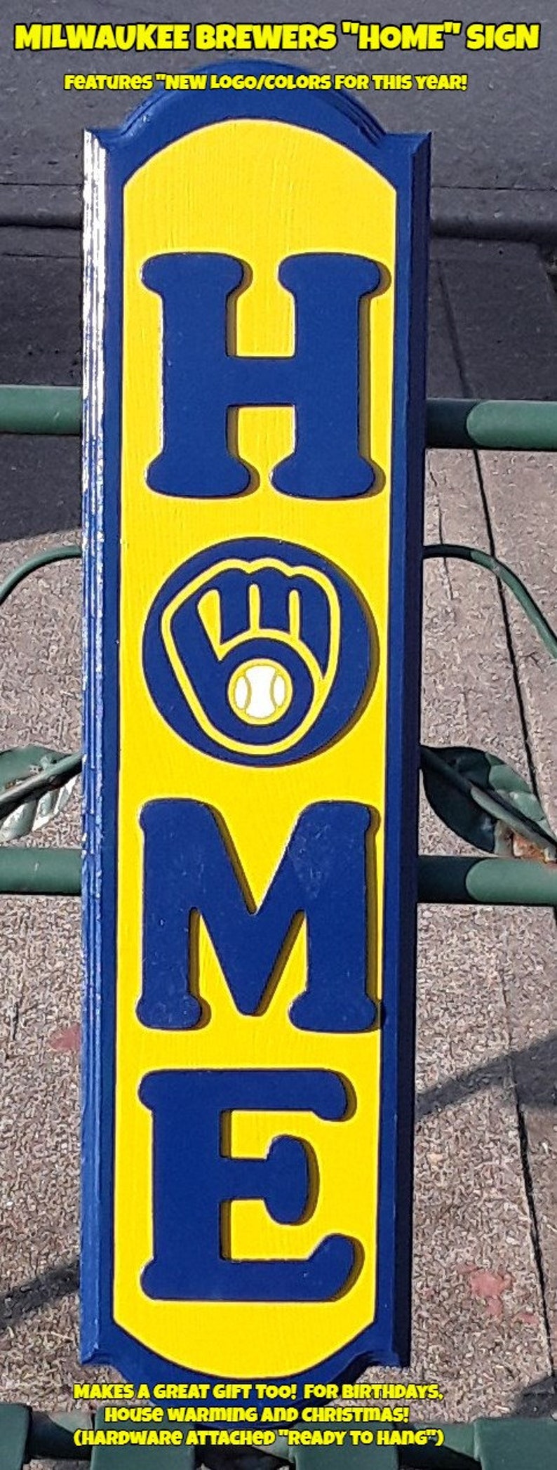 Op de afbeelding: Een houten bord met het woord "HOME" in blauwe letters op een gele achtergrond. De letter "O" is vervangen door het logo van het Milwaukee Brewers honkbalteam, een blauwe handschoen met een witte honkbal. Het bord heeft de tekst "MILWAUKEE BREWERS "HOME" SIGN" bovenaan en "MAKES A GREAT GIFT TOO! FOR BIRTHDAYS, HOUSE WARMING AND CHRISTMAS! (HARDWARE ATTACHED "READY TO HANG")" onderaan.