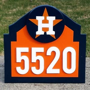 Può includere: Un cartello di numero civico in legno blu e arancione con una stella bianca e la lettera "H" al centro. Il numero civico 5520 è visualizzato in bianco su sfondo arancione.