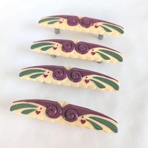 May include: Four curved drawer pulls featuring a cream-colored base with dark purple central swirls, dark green leaf-like accents, and small red heart details. Each handle has two metal mounting posts on the underside for attachment to furniture.