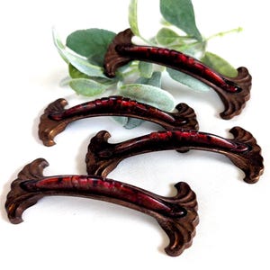 May include: Four antique bronze cabinet pulls with a dark red center. The pulls have a curved design with ornate detailing on the ends. The hardware is likely for a dresser or cabinet.