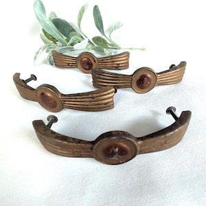 May include: Four antique bronze-colored drawer pulls with a central circular accent. Each pull has a curved, ribbed design and includes a screw for mounting. The pulls are arranged on a white surface with a sprig of green foliage.