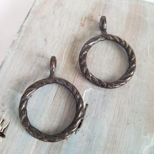 May include: Two bronze-colored curtain rings with a twisted rope design. Each ring has a small loop at the top for hanging. The rings are displayed on a light blue and white wooden surface.