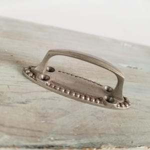 May include: A silver-toned metal handle with an arched grip and an oval base. The base has decorative beading around the edges and four screw holes. The handle is set against a weathered, light-colored wooden surface.