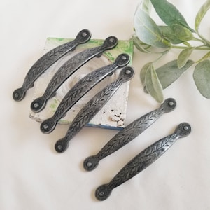 May include: Set of six antique-style cabinet handles. Each handle is a dark gray color with a leaf design. The handles are curved with rounded ends and are approximately 5 inches long.