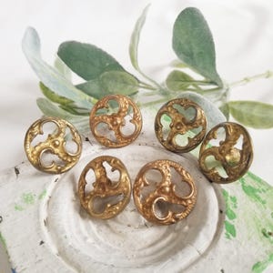 May include: A set of five circular brass buttons, each featuring an intricate openwork design with three symmetrical, swirling scroll-like motifs radiating from a central point. The metal has a warm, aged gold-toned finish with visible texture.