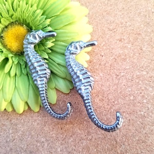 May include: Two silver seahorse-shaped hooks. The hooks are detailed with textured scales and curved tails. They rest on a light brown surface next to a green and yellow flower, creating a coastal or nautical theme.