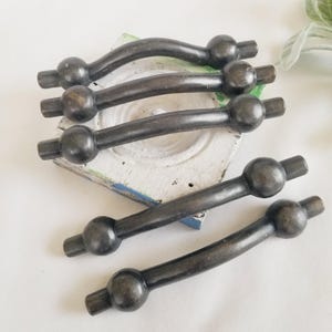 May include: Five dark metallic cabinet pulls with a curved design and spherical accents at each end. Each pull features a smooth, matte finish with a slightly weathered appearance, showcasing a consistent industrial aesthetic across all five pieces.