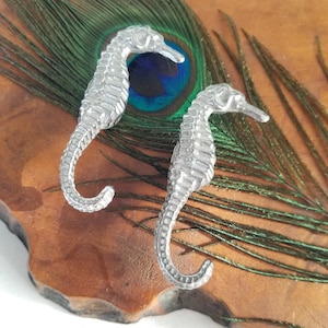 May include: Two silver seahorse figurines. The seahorses are detailed with textured surfaces and curved tails. They rest on a wooden surface with a peacock feather in the background. The seahorses are approximately 4 inches in length.