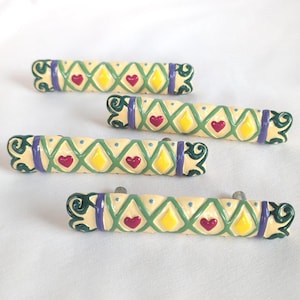 May include: Four cylindrical drawer pulls with a cream-colored base, featuring a green lattice pattern, purple bands, and alternating yellow diamond and red heart motifs. Each pull has dark green scrollwork at the ends and small blue dots.