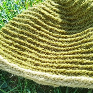 Puede incluir: Un gorro tejido a mano de color verde oliva con un borde beige claro. El gorro está hecho de hilo grueso y tiene un diseño texturizado y acanalado. El gorro está sobre hierba verde.