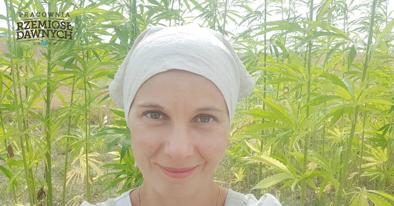 May include: A woman wearing a light gray head covering stands in a field of green hemp plants. The text "PRACOWNIA RZEMIOSŁ DAWNYCH" is visible in the upper left corner of the image.