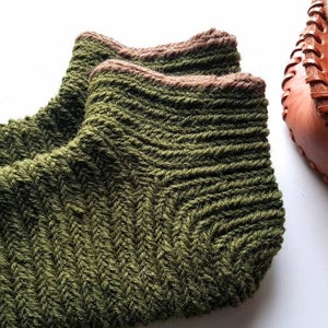 Può includere: Un paio di calzini verdi lavorati a mano con un motivo strutturato e bordi marroni. Sullo sfondo, una borsa in pelle marrone con cuciture decorative.