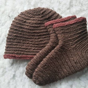 Puede incluir: Un gorro de punto marrón y un par de calcetines a juego. El gorro y los calcetines tienen una textura acanalada con un ribete marrón rojizo. Los artículos se muestran sobre una superficie blanca y esponjosa.